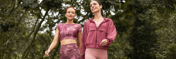 Duas mulheres caminhando em uma trilha arborizada, usando conjuntos esportivos em tons de rosa